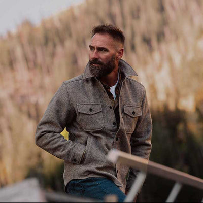 Brown wool Shirt jacket, textured brown overshirt and blue jeans, standing outdoors in front of a soft-focus background of dry grass and trees.
