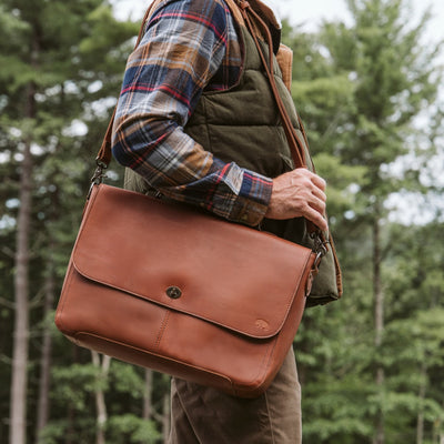 Man outdoors wearing a plaid shirt and vest, carrying a stylish brown leather messenger bag over his shoulder in the forest.