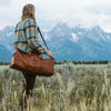 Adventurer in plaid carrying a brown leather bag through a scenic meadow in Jackson Hole, Wyoming, under cloudy mountain skies.
