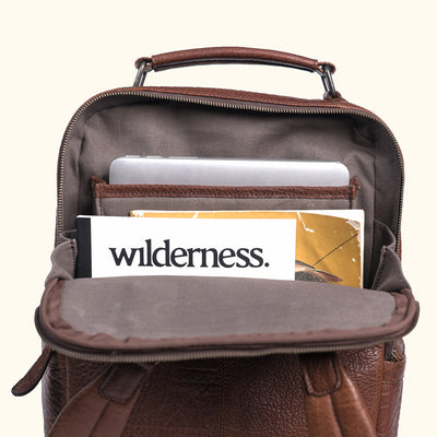 Functional brown leather backpack interior view, showcasing compartments with a tablet, phone in case, books, and travel postcard.