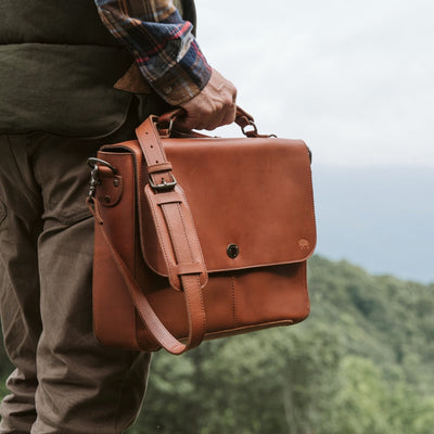 Man outdoors holding a luxurious autumn brown leather messenger bag with robust straps and hardware, against a scenic forest backdrop