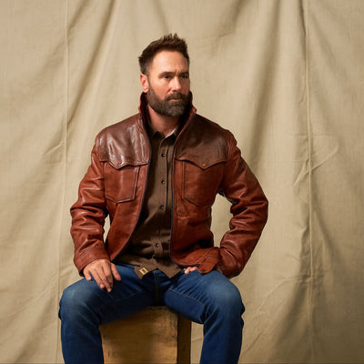 Man wearing a brown leather jacket sitting on a stool against a beige curtain.