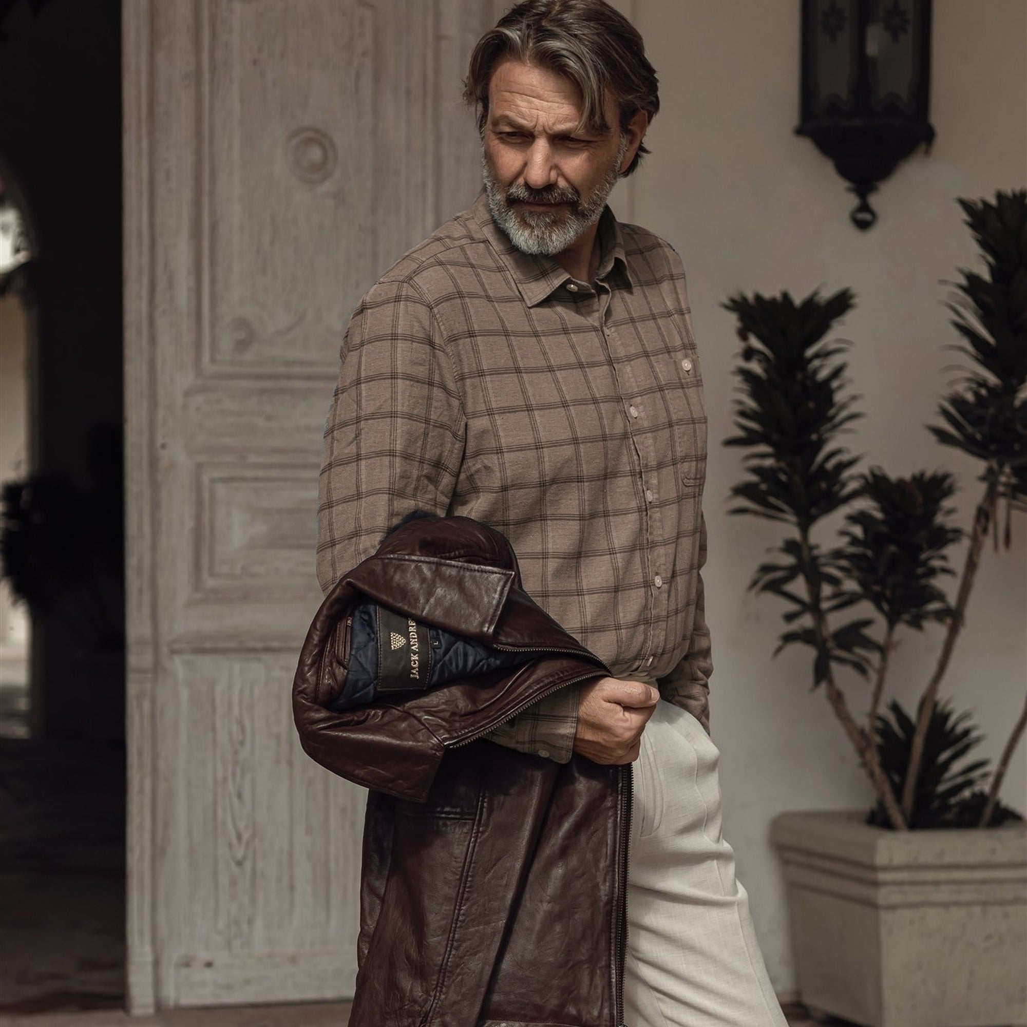 Man in the Windsor Cotton Shirt in Timber Pane carrying a leather jacket through a courtyard