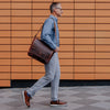 Sleek dark oak leather satchel messenger bag carried by a man in urban attire, featuring a polished finish and spacious design.
