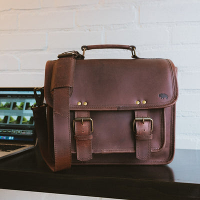 Luxurious dark oak leather camera bag with sturdy handle, detachable shoulder strap, and brass hardware.