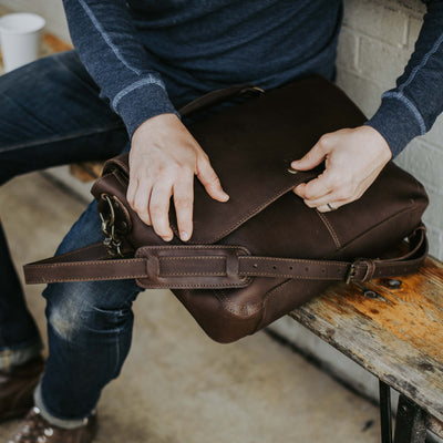 Man seated at a rustic outdoor table, handling a dark brown leather messenger bag with an adjustable strap and secure stitching details.