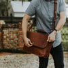 Man in casual attire adjusting a stylish brown leather messenger bag with adjustable strap and secure flap closure outdoors.