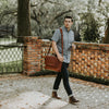 Young man walking in a scenic outdoor setting, stylishly carrying a brown leather messenger bag with a cross-body strap.