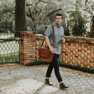 Young man walking in a scenic outdoor setting, stylishly carrying a brown leather messenger bag with a cross-body strap.
