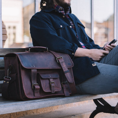 Distressed Dark Oak Leather Briefcase with sturdy handle and shoulder strap, accompanying a man on a public bench.