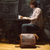 Richly toned vintage leather satchel messenger bag placed next to a casually dressed man in a coffee shop, highlighting its elegant and functional design.