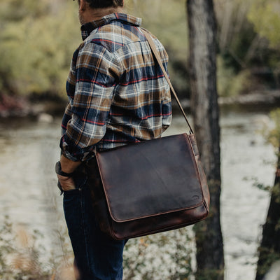 Man in a plaid shirt and jeans overlooking a river, carrying a sleek dark brown leather messenger bag, immersed in nature.