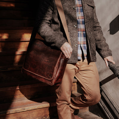 Stylish man descending stair in a coat and khakis, carrying a vintage dark brown leather messenger bag in sunlight.