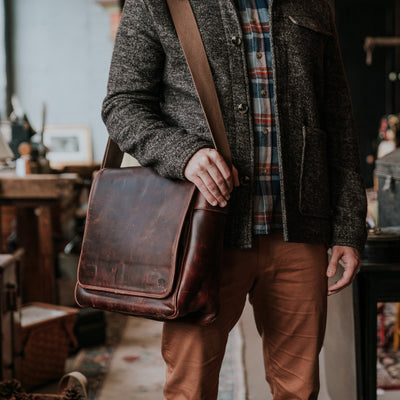 Man in a wool coat and plaid shirt holding a distressed dark brown leather messenger bag, standing in a cozy vintage workspace.