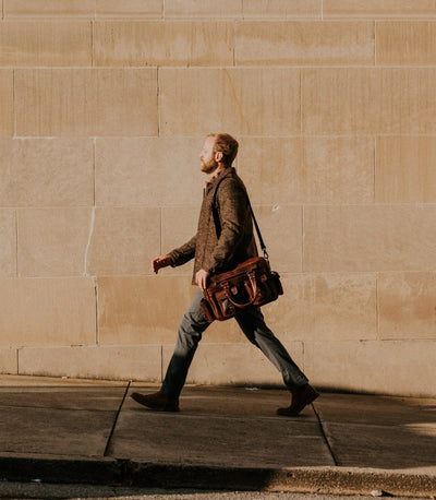 Man walking confidently with a rugged dark oak leather pilot bag featuring durable straps, spacious compartments, and distinctive buckle closures. Perfect for stylish travelers.