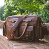 Men's dark oak leather pilot bag sitting on a brick ledge, showcasing its vintage style with multiple pockets, sturdy handles, and a broad strap for comfort.