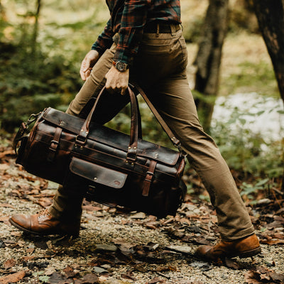 Man walks through woodland trail carrying a dark leather duffle bag with brass buckles and sturdy handles.