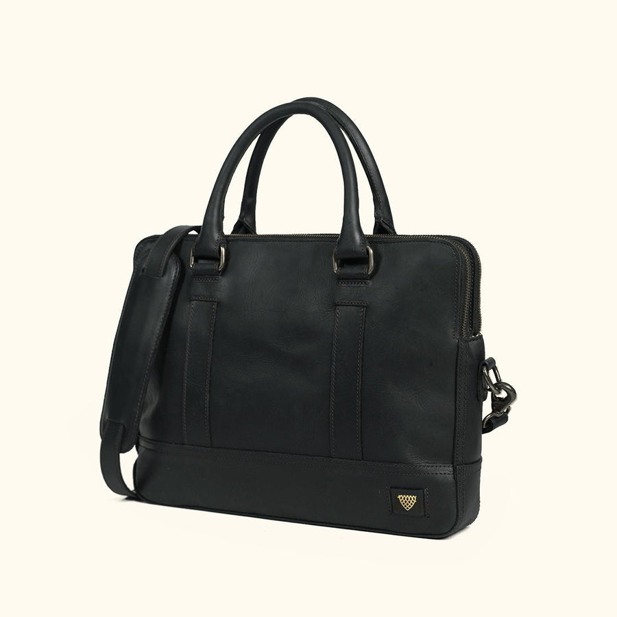 A black leather briefcase with dual top handles, a detachable shoulder strap, and antique nickel hardware, with a small gold shield logo on the bottom right corner, set against a solid cream background.