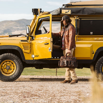 A man in a brown leather jacket and beige pants stands beside a bright yellow off-road safari vehicle, Land Rover Wolf, holding a brown leather briefcase in an outdoor desert setting.