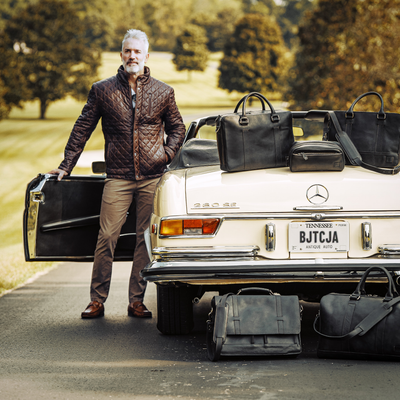 A man in a quilted leather jacket stands confidently by a vintage car, surrounded by luxury leather bags.