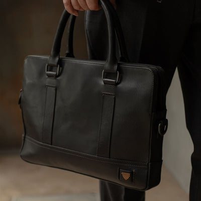 A black leather briefcase with top handles and antique nickel hardware, carried by a person wearing dark clothing. A small gold shield logo patch is visible near the lower right corner.