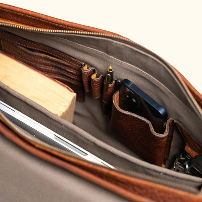 Interior view of a brown leather messenger bag showing organized pockets holding a laptop, phone, pen, and notebook.