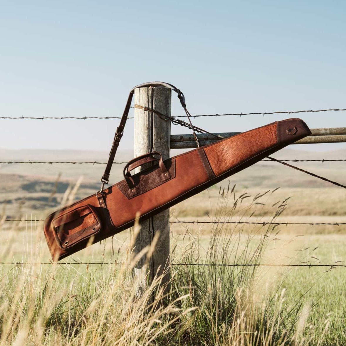 Full grain leather shotgun case resting across a wire fence in a field of dry autumn grass