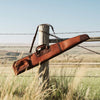 Rustic leather shotgun case draped over a barbed wire fence, with a backdrop of open fields.