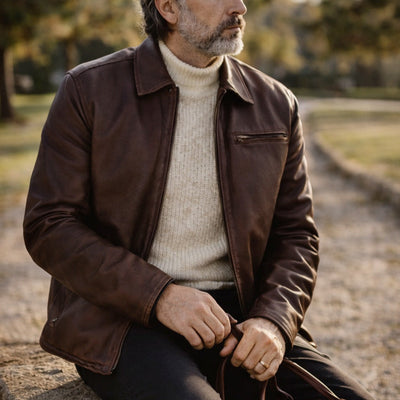 Man in a brown leather jacket and white sweater sitting on a stone wall with a blurred natural background
