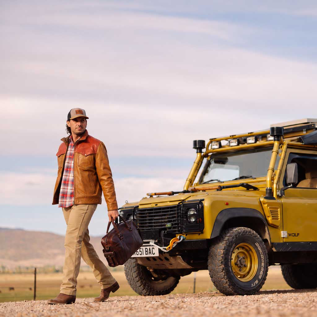 A man in a brown leather jacket and beige pants walks beside a yellow off-road Land Rover camp vehicle with a black canvas roof tent in an open desert landscape under a blue sky.