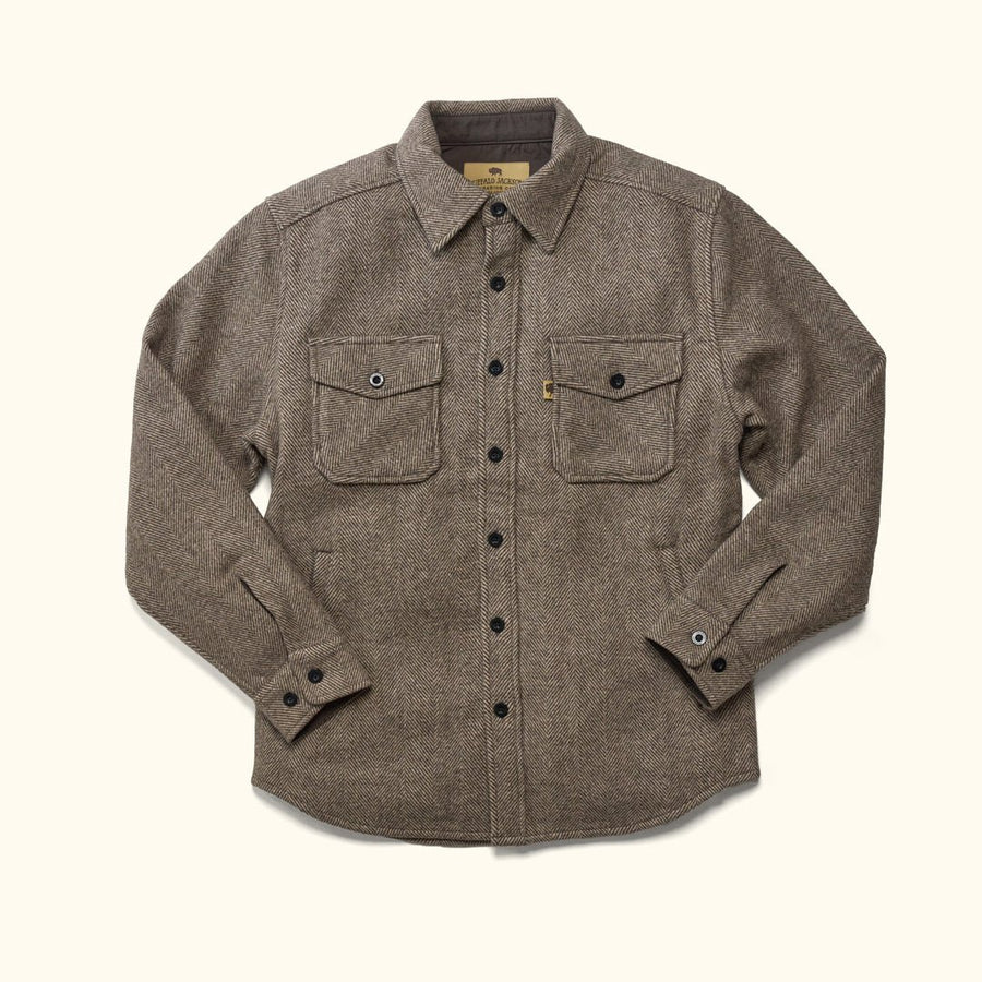 Brown wool Shirt jacket,  textured brown overshirt and blue jeans, standing outdoors in front of a soft-focus background of dry grass and trees.