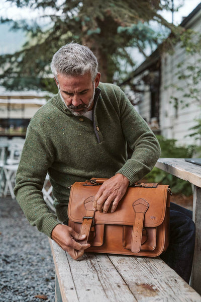 Mature man in green sweater using Roosevelt amber brown leather briefcase on a rustic wooden table.