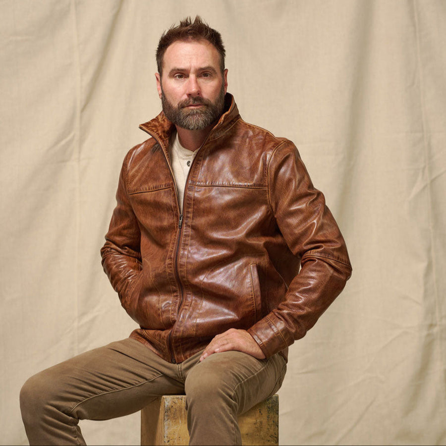 Man wearing a brown leather jacket sitting on a wooden stool against a beige background