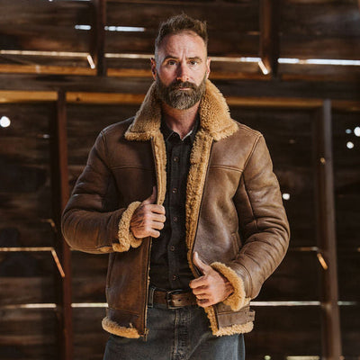 A bearded man wearing a brown shearling-lined leather jacket stands inside a rustic wooden barn, holding the front of the jacket open.