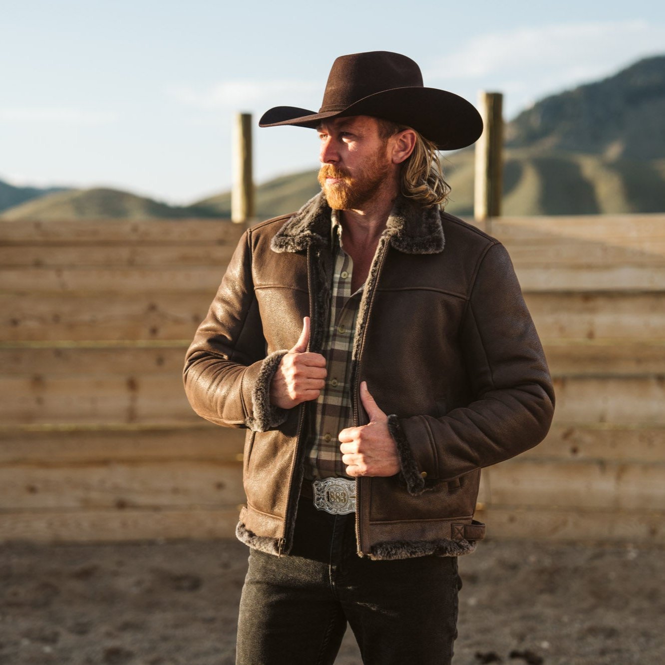 Cowboy in a hat wearing the Shearling Leather Bomber Jacket in Espresso Brown by a wooden corral