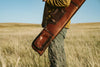 Man carrying a leather shotgun case slung over his shoulder, standing in a scenic rural landscape.