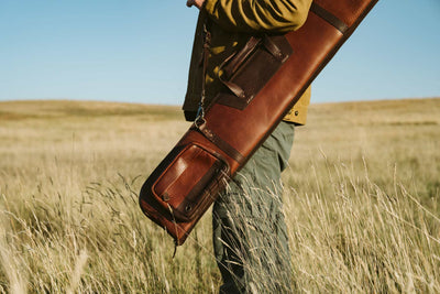 Man carrying a leather shotgun case slung over his shoulder, standing in a scenic rural landscape.