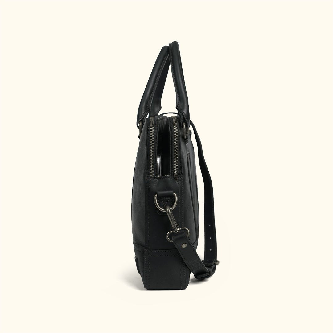 Side view of a black leather attaché bag with antique nickel zippers and hardware on a cream background