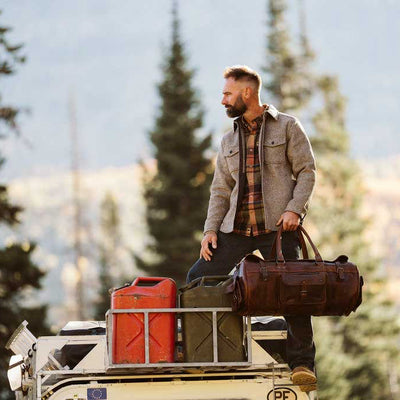 A man stands on the back of a vehicle in a forested outdoor setting, holding a brown duffel bag. He is wearing a brown overshirt over a plaid shirt and dark pants, with a red fuel canister and other bags strapped to the vehicle rack.