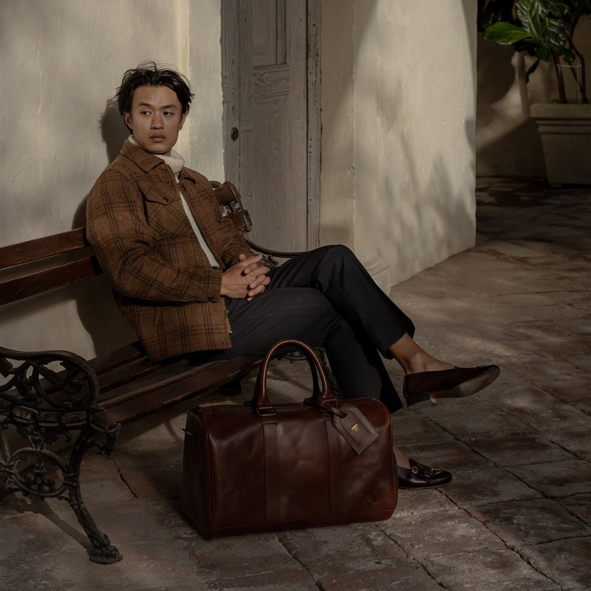 Man in the Aberdeen Wool Jacket in Dunbar Thicket seated on a bench with a leather duffle