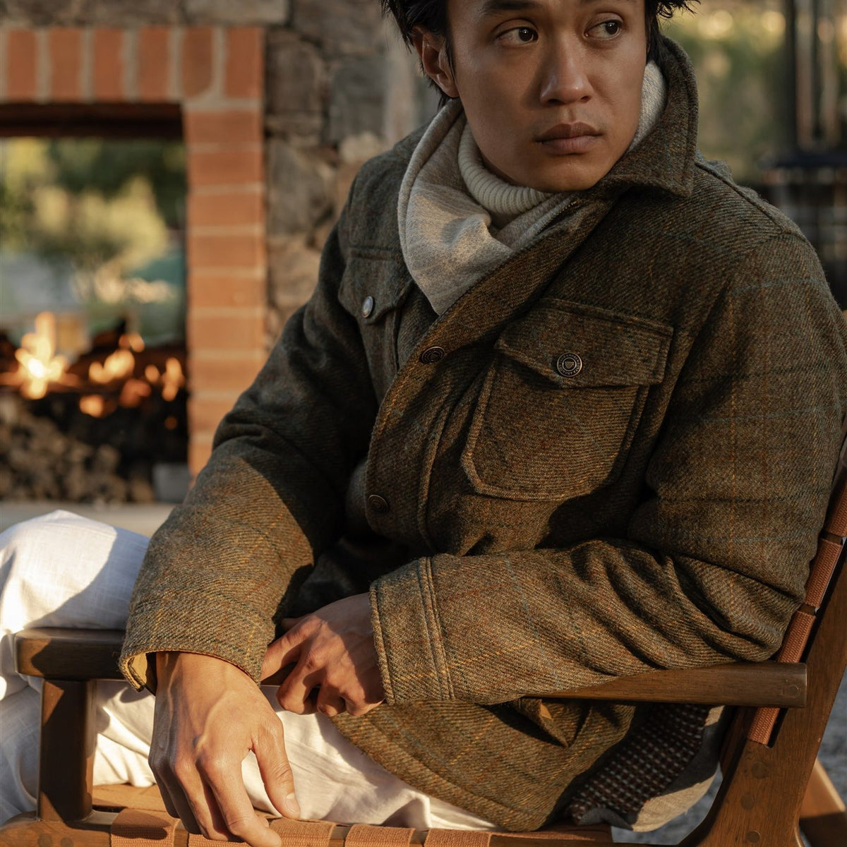 Man in the Aberdeen Wool Jacket in Fir Heather and a gray scarf seated by an outdoor fire