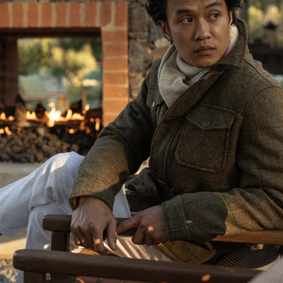 Man in the Aberdeen Wool Jacket in Fir Heather seated on a wooden bench by an outdoor fire