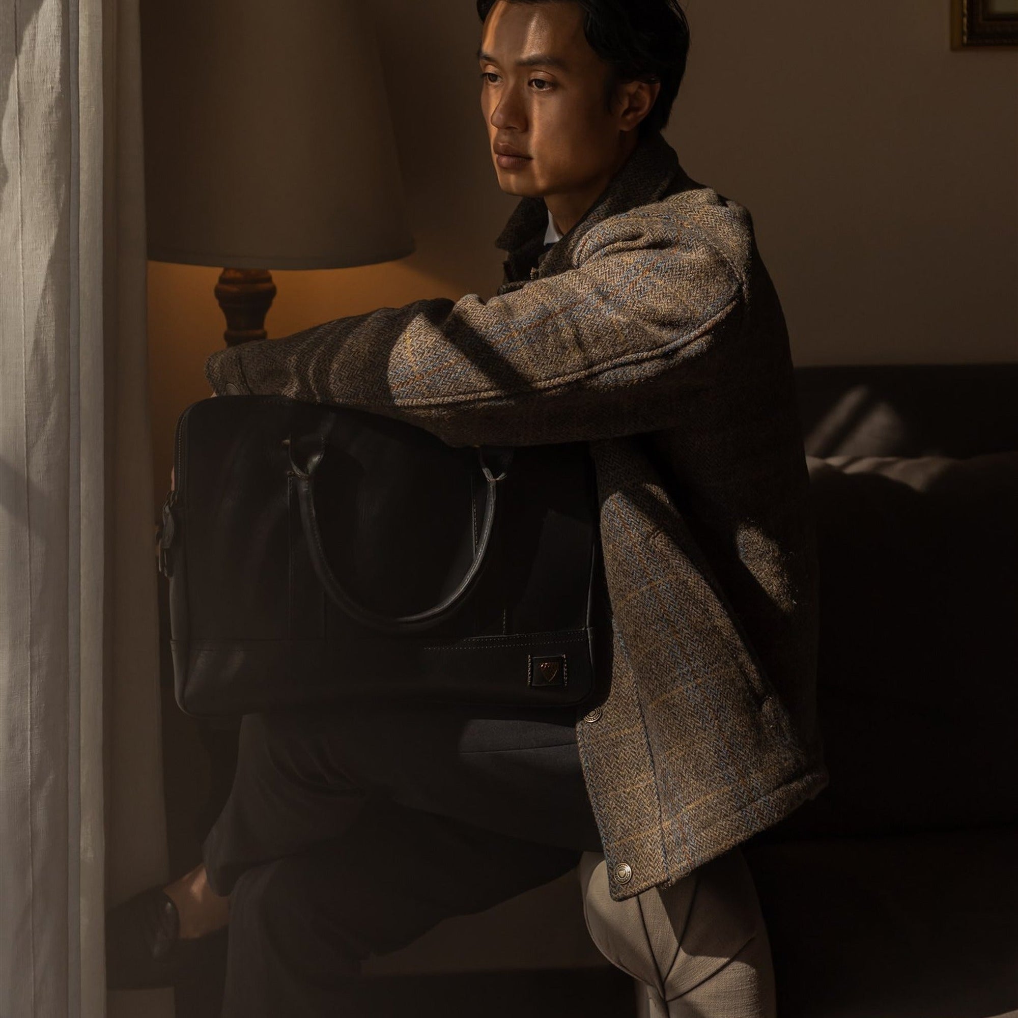 Man in the Aberdeen Wool Jacket in Thistle Bark Plaid seated in an armchair with a leather bag