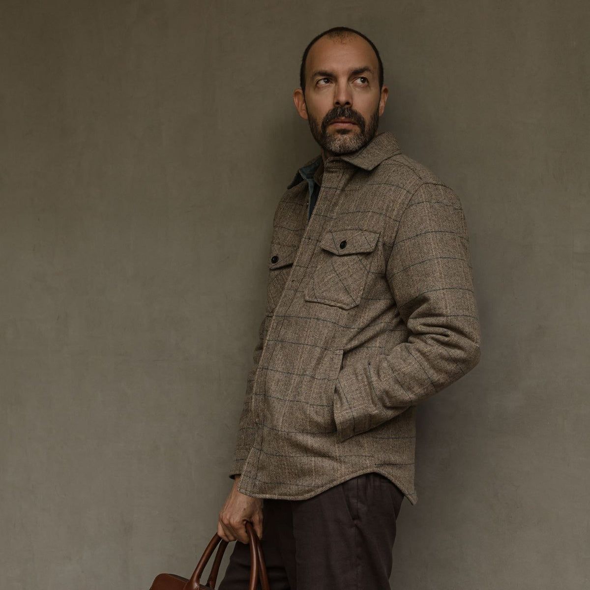 Man in the Aberdeen Wool Twill Shirt Jac in Thistle Grove leaning on a wall with a leather briefcase