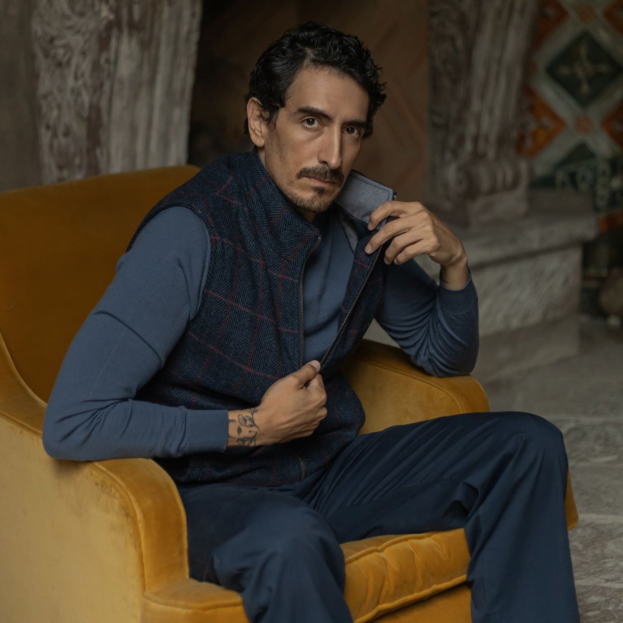 Man in the Ashton Wool Herringbone Vest in Clan Blue seated in a mustard velvet armchair