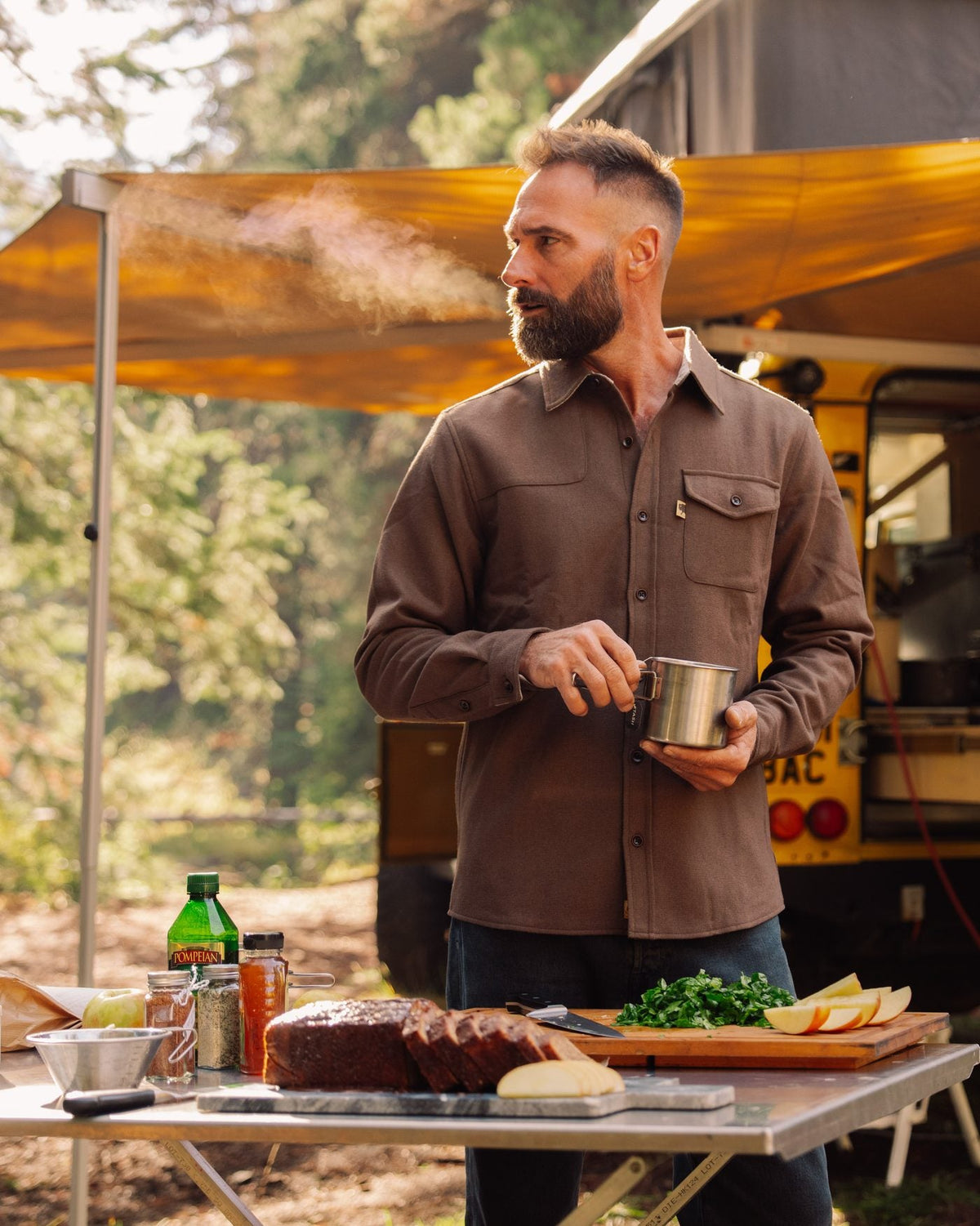Man in a brown button-up shirt holding a camp mug under an awning at a campsite