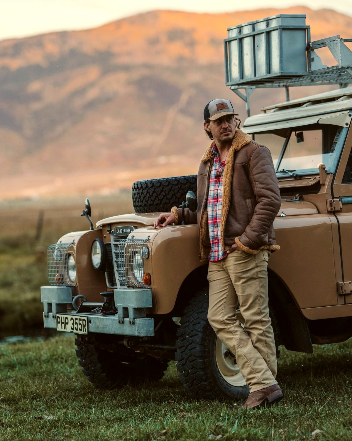 Man in a shearling-collar leather jacket leaning on a vintage Land Rover below mountains at dusk