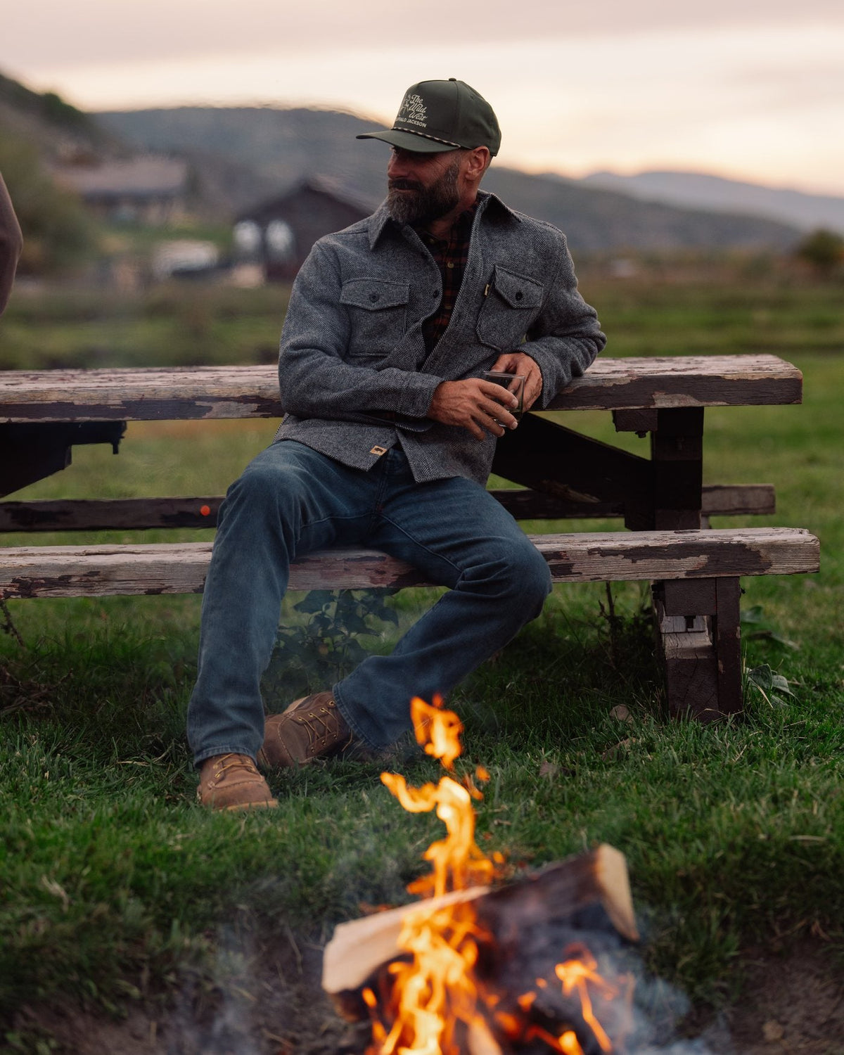 Man in a gray herringbone wool shirt jac sitting by a campfire at dusk