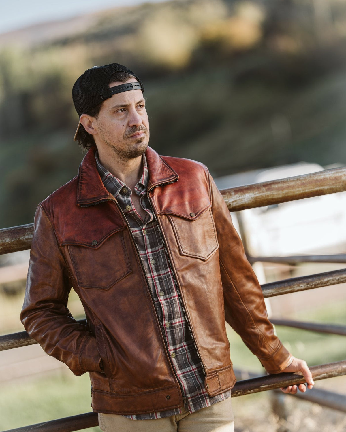 Man in a Western leather jacket over a plaid flannel leaning on a ranch fence