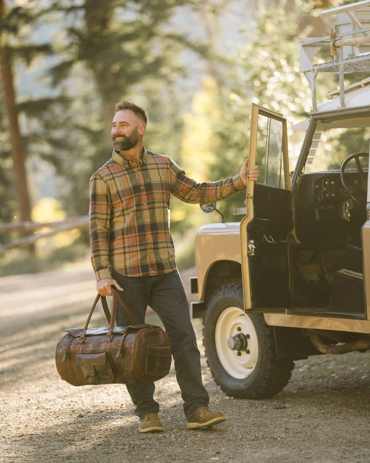 Man in a plaid flannel shirt carrying a leather duffle beside a vintage Land Rover